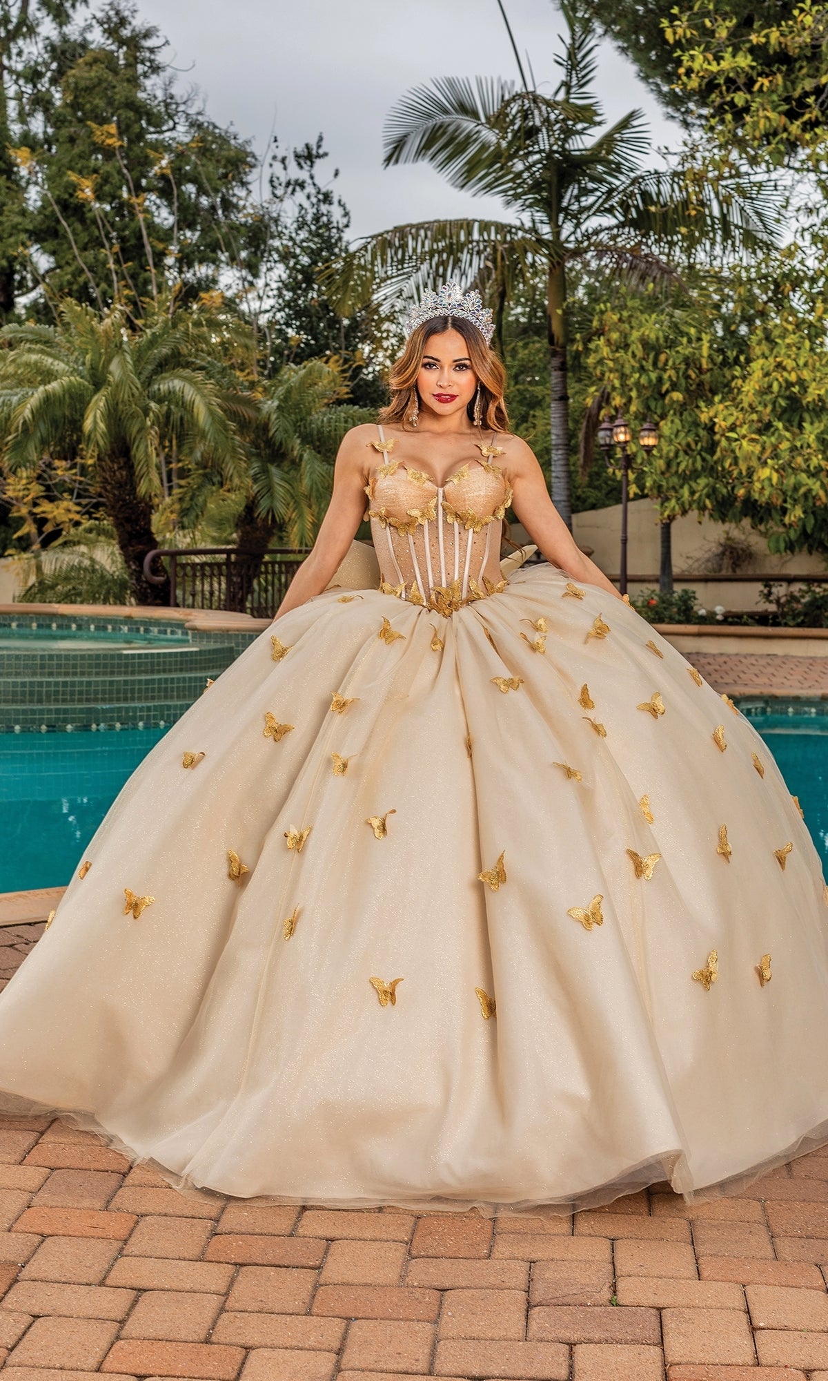 A woman stands by a pool in the Quinceanera Dress 1815 By Dancing Queen, featuring gold fabric, butterfly embellishments, a sheer corset bodice, and sweetheart neckline. Palm trees and lush greenery frame the elegant scene.